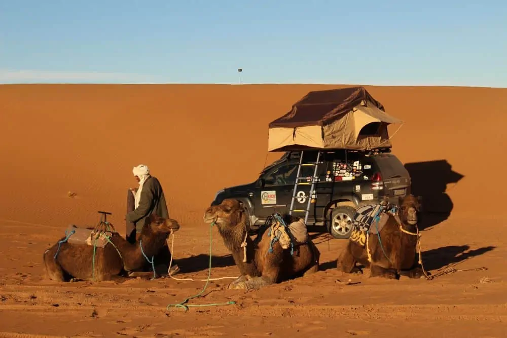 THE ULTIMATE ITINERARY FOR AN UNFORGETTABLE MOROCCO ROAD TRIP featured by top international travel blogger, The Common Traveler; Image: SUV with tent in Morocco dessert with camels in foreground