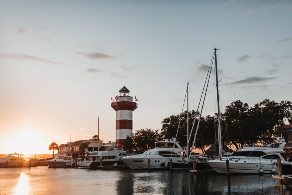 image: boats at sunrise with lighthouse at Hilton Head Island, SC | Best Places to Visit in South Carolina: image of a red and white stripe light house and sailboats in Hilton head Island. 